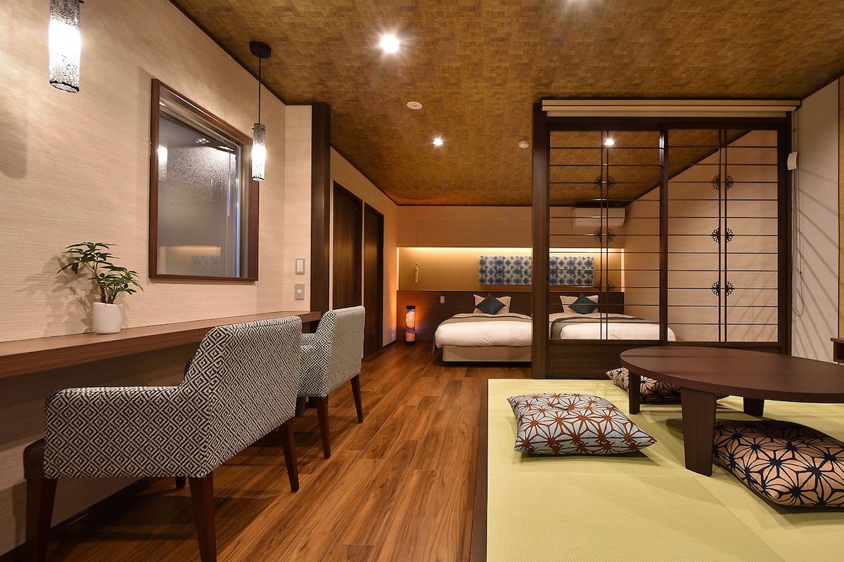 A hotel room with a wooden floor, a bed with a white headboard, a TV on the wall, a desk with a chair, and a sliding door.