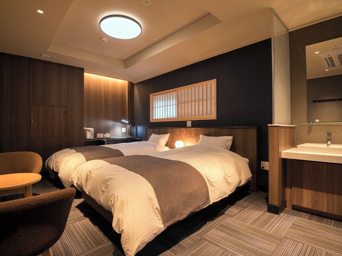 A hotel room with two beds, a sitting area, a bathroom with a mirror and sink, and a ceiling light.