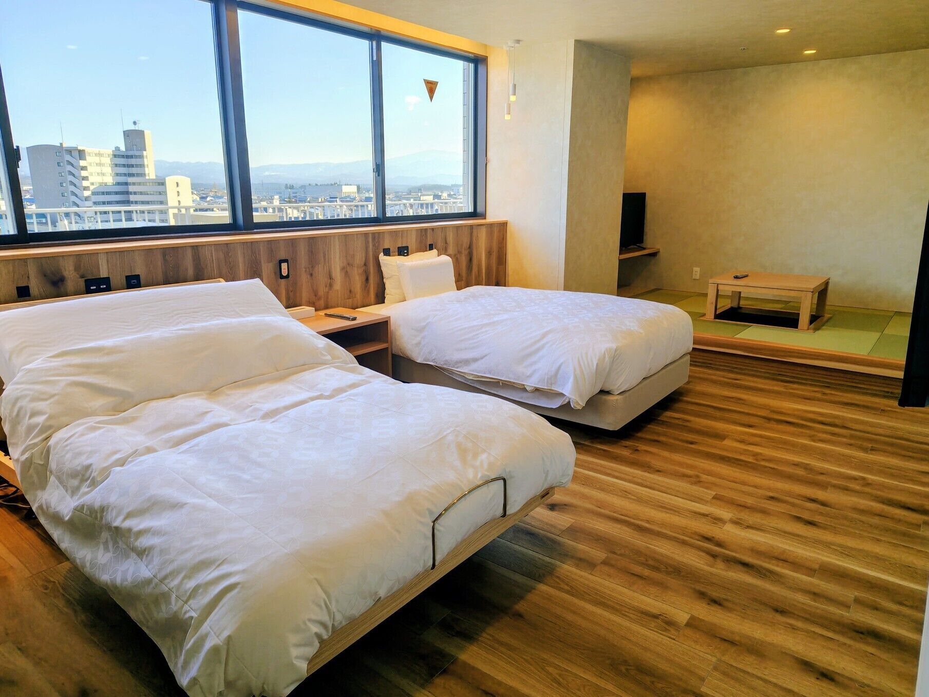 A hotel room with two beds, a wooden floor, a large window with a city view, and a small table.
