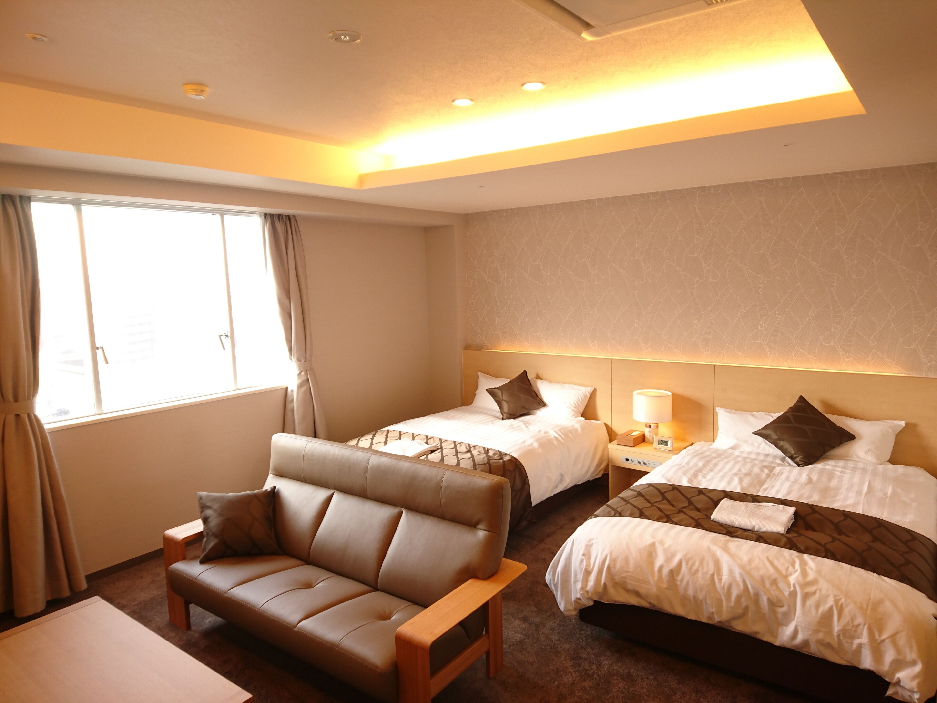 Quarto de hotel com duas camas, um sofá, uma mesa de cabeceira com luminária e uma janela com cortinas.