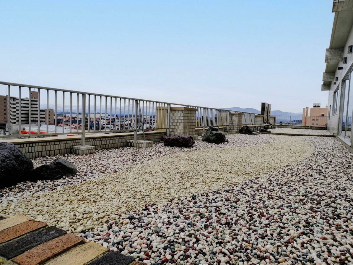 A rooftop terrace with a gravel surface, a metal railing, and a view of the cityscape.