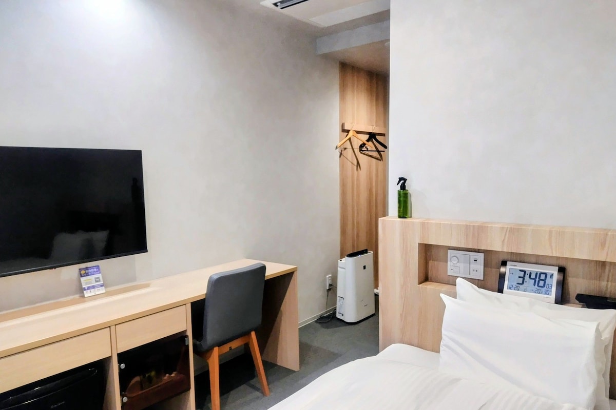 A hotel room with a bed, a desk with a chair, a television, a heater, and a clock.