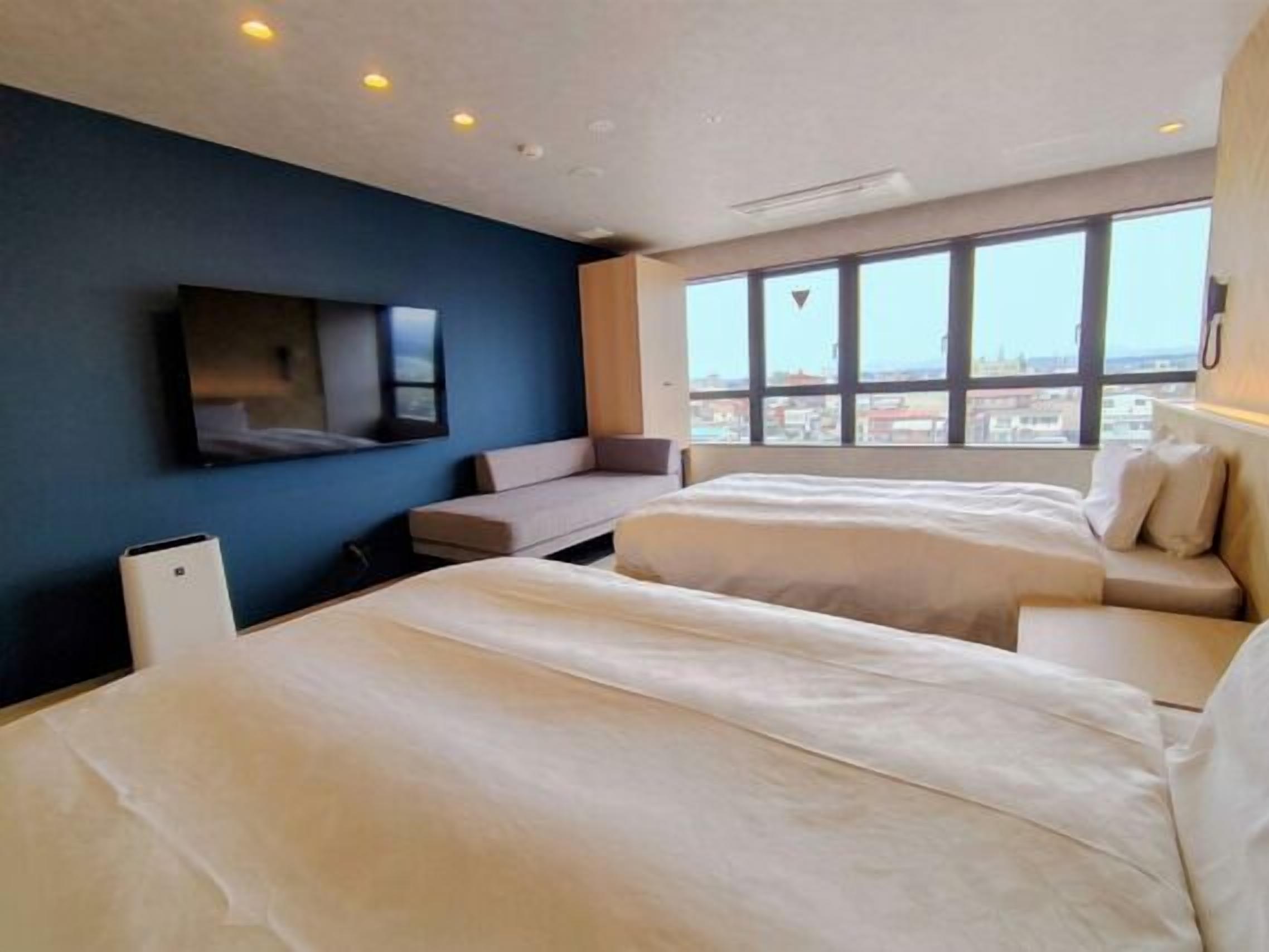 A hotel room with two beds, a TV, a window with a city view, and a small seating area.