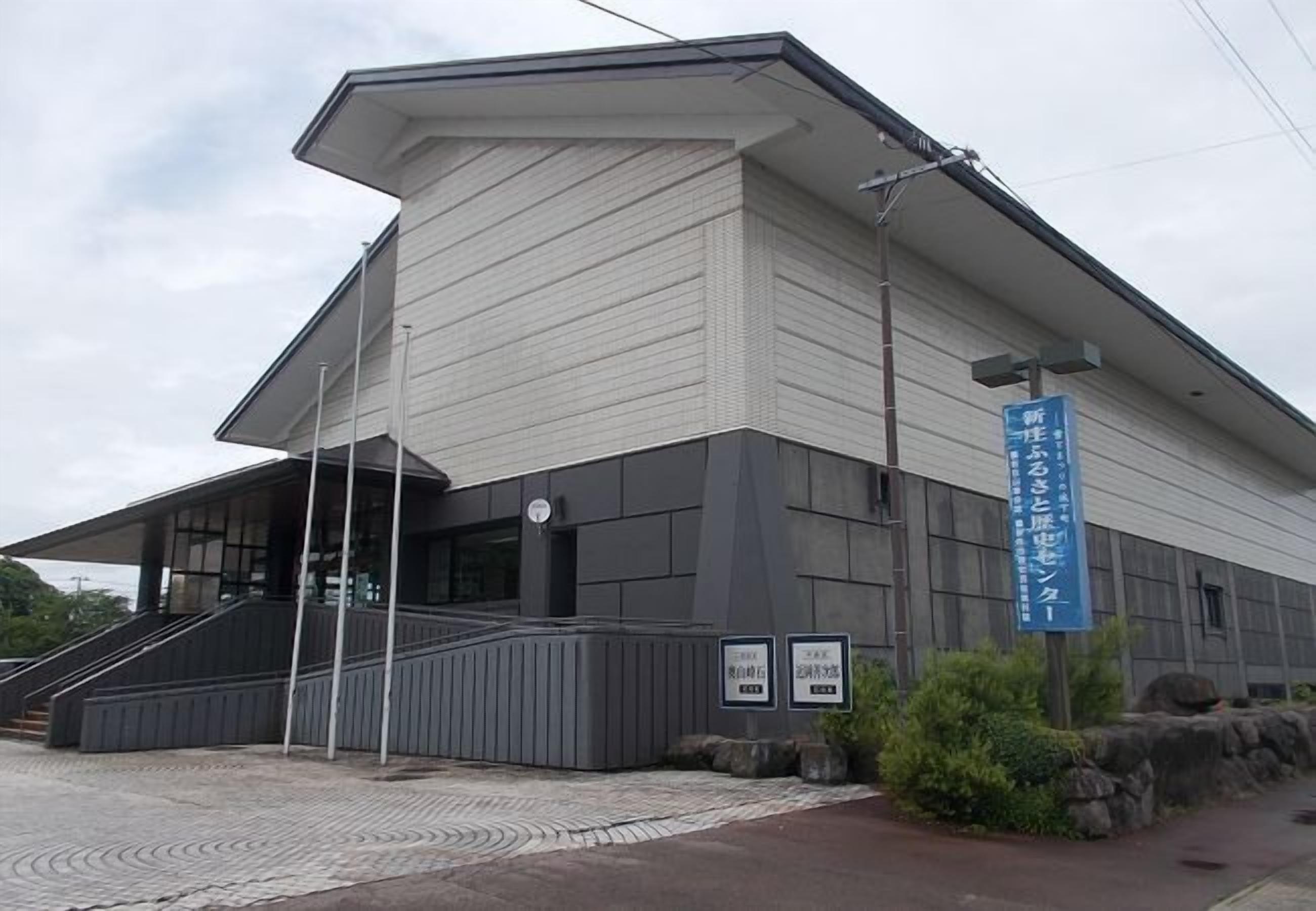 Um edifício moderno com telhado inclinado, uma escadaria que leva à entrada e uma placa azul com texto em japonês.