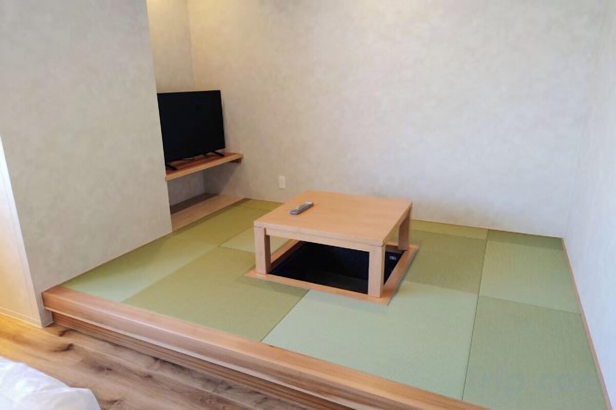 Una habitación con suelo de tatami, un televisor sobre un soporte y una pequeña mesa de madera.