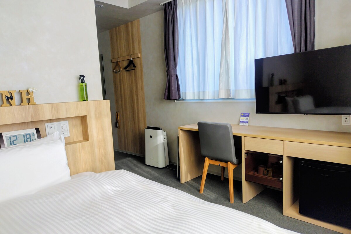 Habitación de hotel con cama, escritorio con silla, televisor, armario y ventana con cortinas.