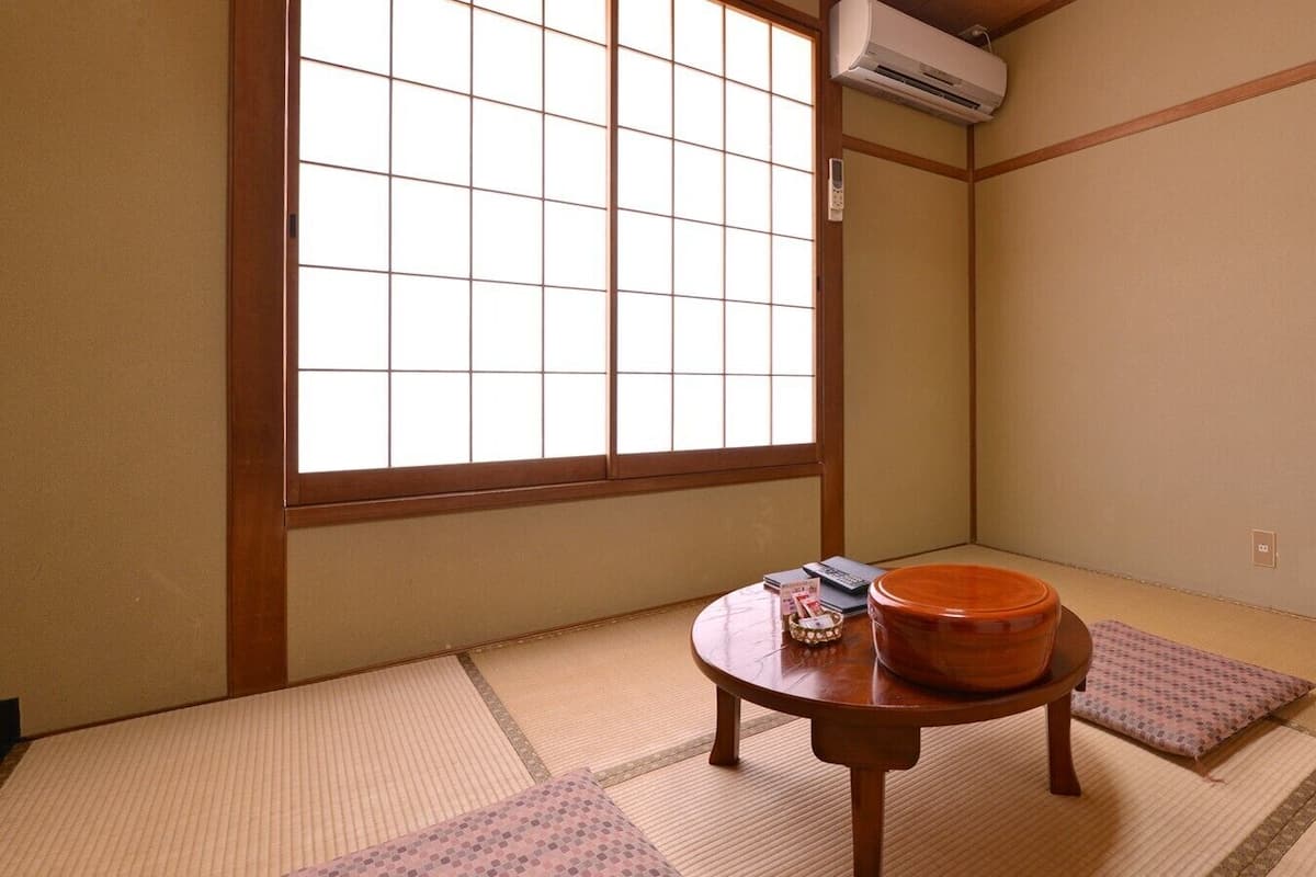 A traditional Japanese room with tatami flooring, a tatami mat table, a wooden table with a bowl, and a window with a grid pattern.