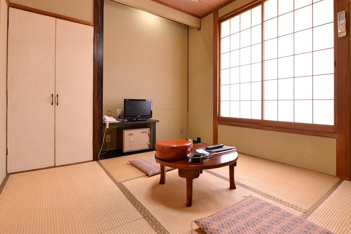 A traditional Japanese room with tatami flooring, a sliding door, a television, a small table with a tea set, and a window with a view.