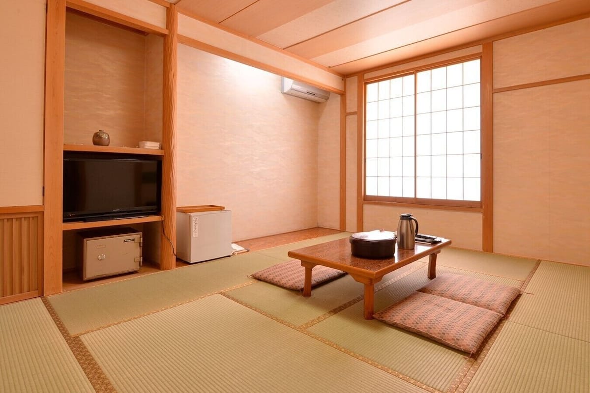A traditional Japanese room with tatami flooring, a television, a small refrigerator, and a window with a view.