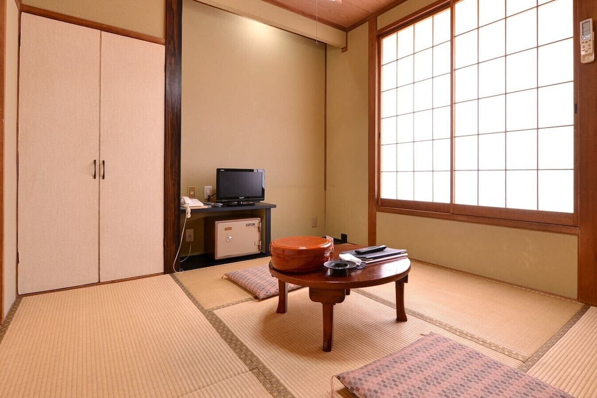 A traditional Japanese room with tatami flooring, a sliding door, a television, a small table with a tea set, and a window with a view.