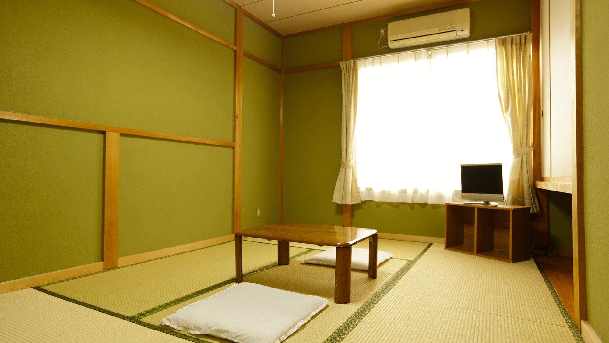 Une pièce de style japonais traditionnel, avec un sol recouvert de tatamis, une petite table en bois, un téléviseur posé sur un meuble et une fenêtre avec des rideaux.
