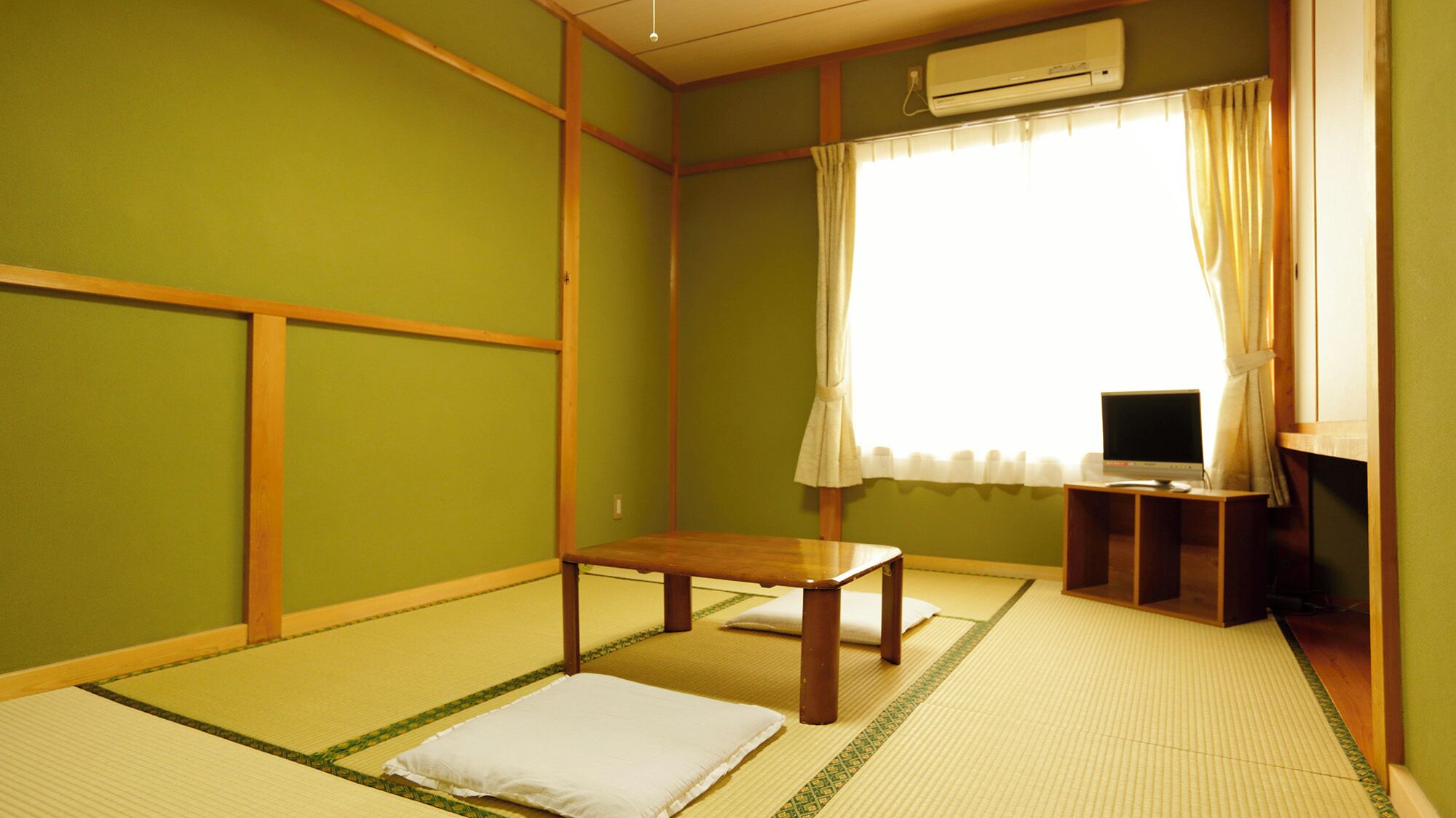 Une pièce avec un sol recouvert de tatamis, un futon servant de lit, une petite table en bois, un téléviseur posé sur un meuble et un appareil de climatisation.