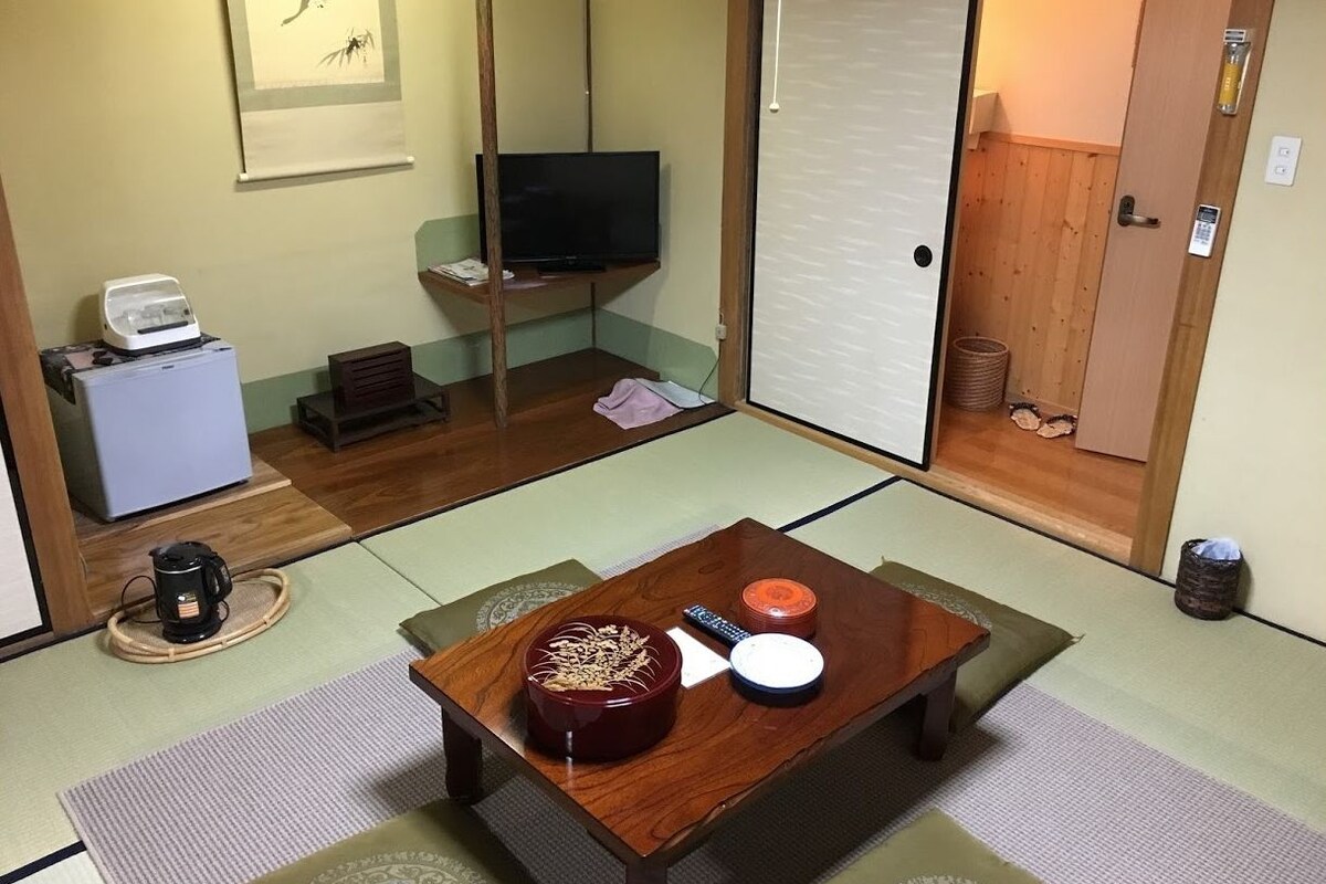 A traditional Japanese room with tatami flooring, a low wooden table with a bowl and a remote control, a television on a stand, and a sliding door.
