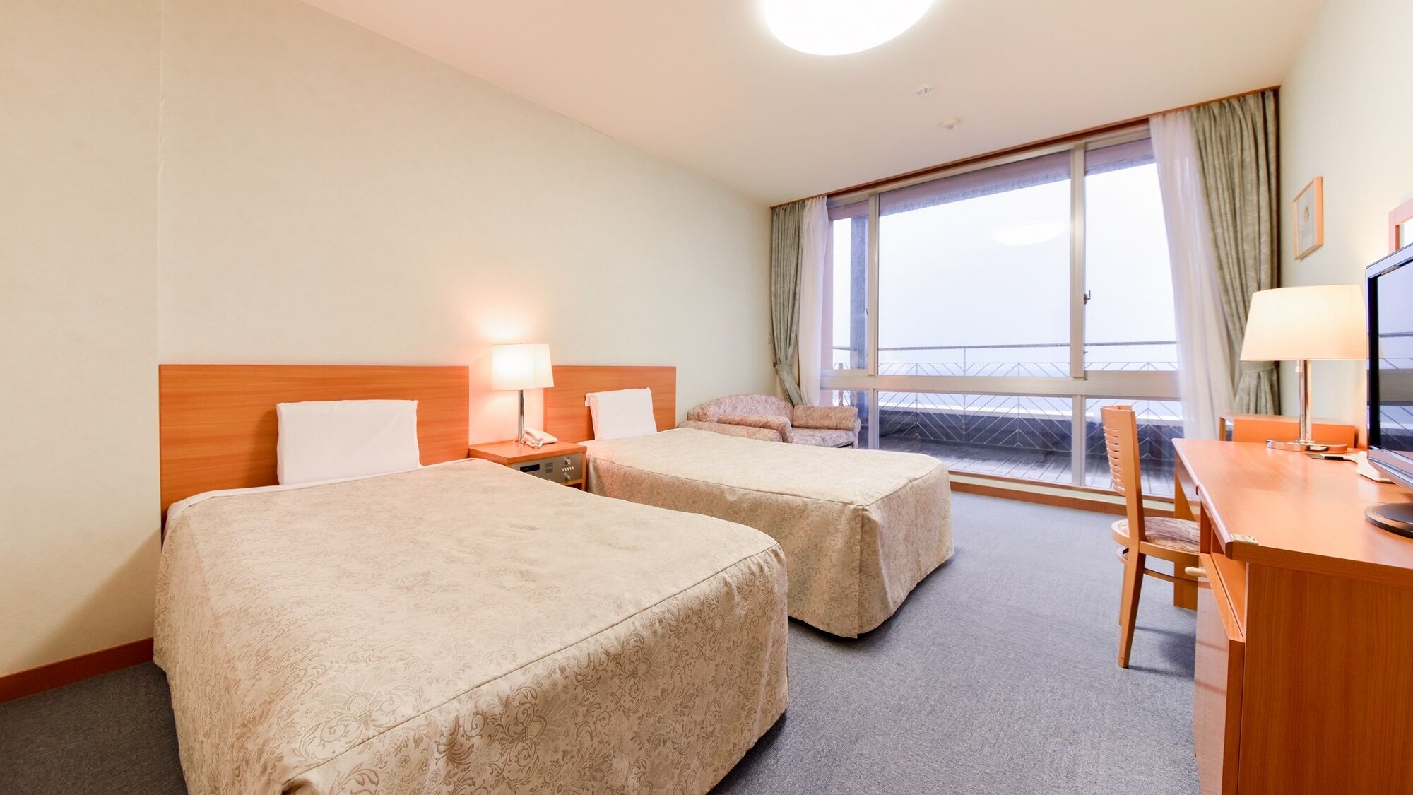 Camera d'albergo con due letti, una scrivania, una sedia, una televisione e un'ampia finestra con tende.