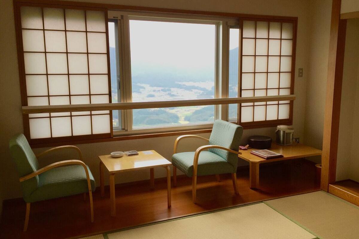 A room with tatami flooring, a low table, two chairs, and a large window with a view of a landscape.