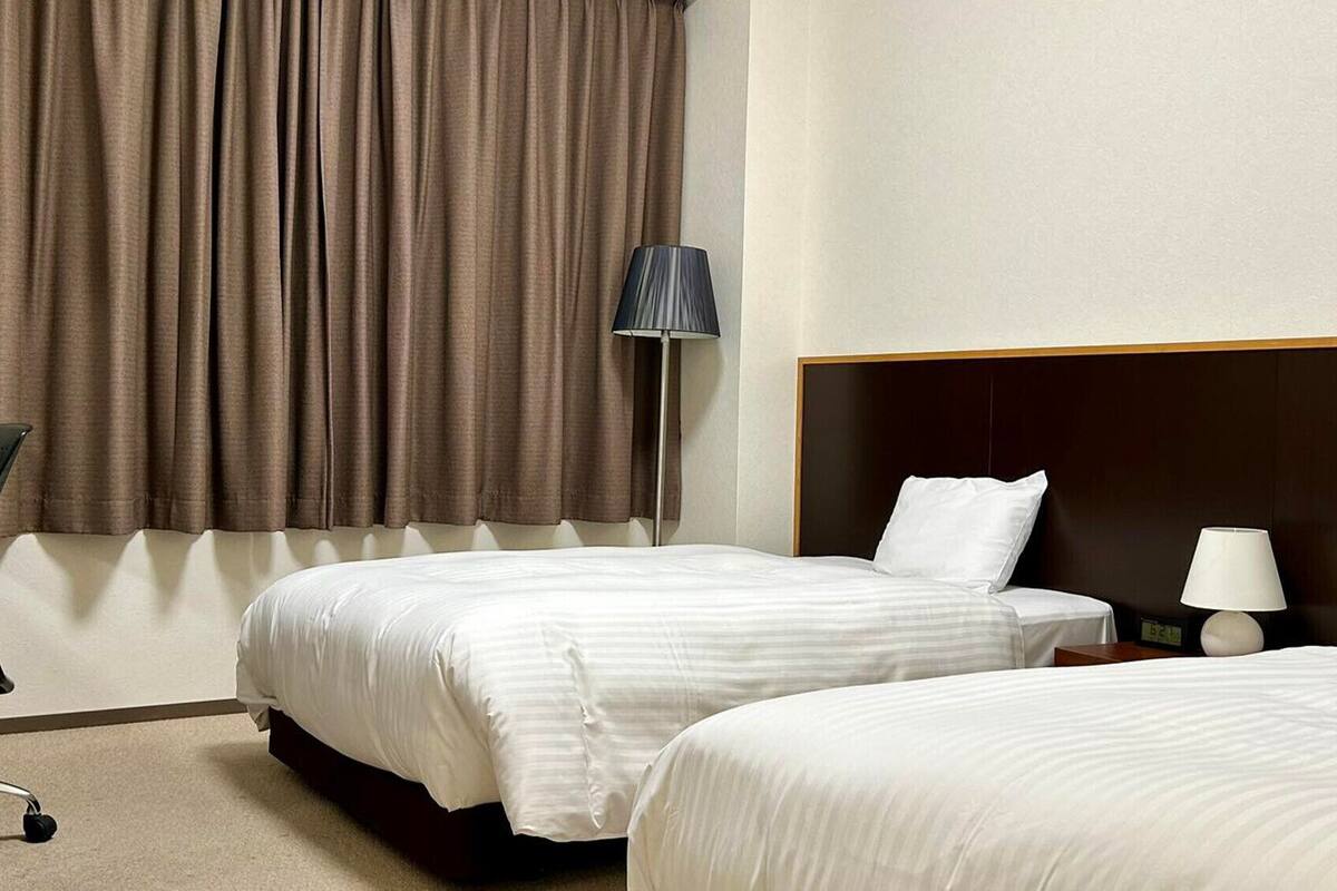 A hotel room with two beds, a desk, a chair, and a lamp.