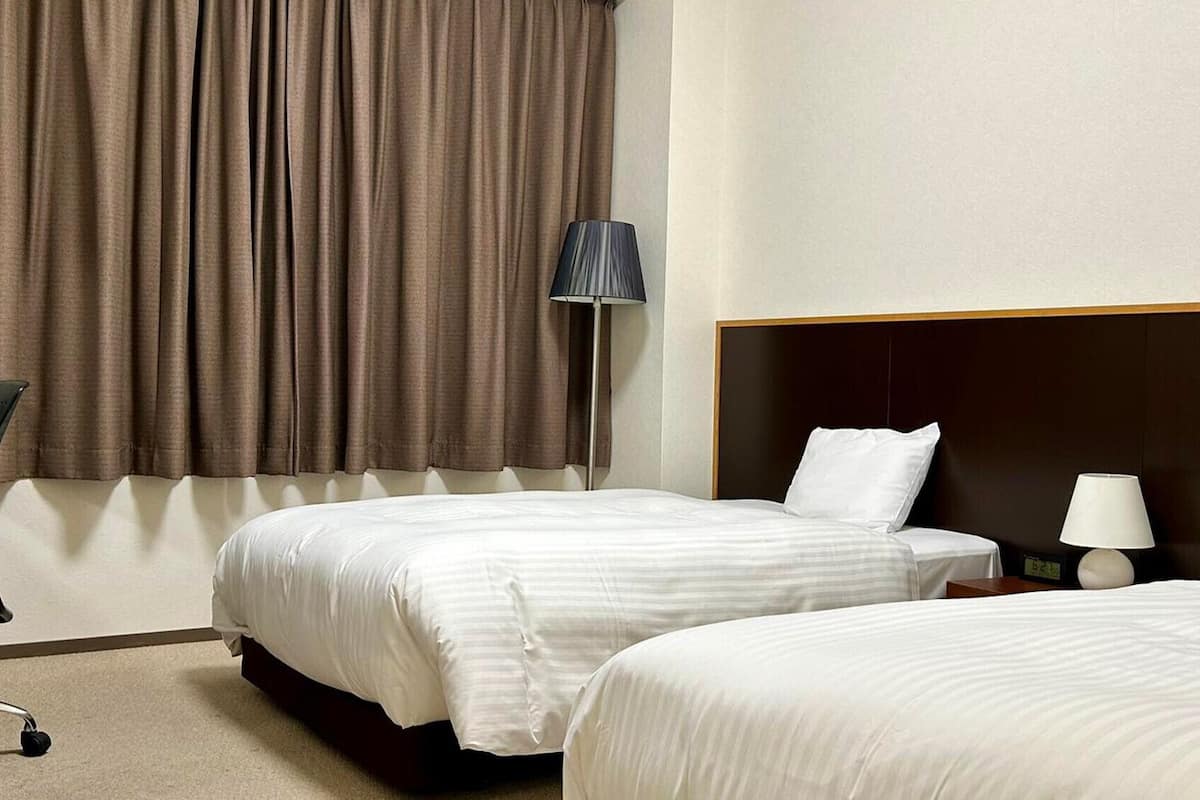 A hotel room with two beds, a desk, a chair, and a lamp.