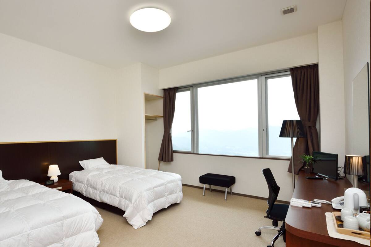A hotel room with two beds, a desk with a chair, a window with curtains, and a view of the sky.