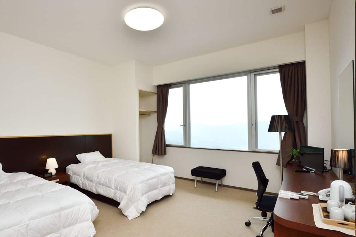 A hotel room with two beds, a desk with a chair, a window with curtains, and a view of the sky.