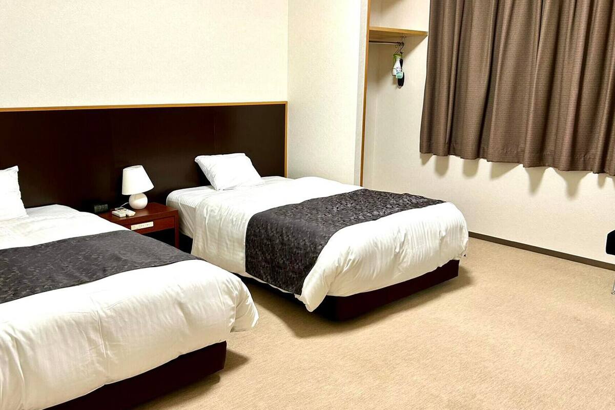 A hotel room with two beds, a nightstand, a lamp, a wardrobe, and a window with curtains.