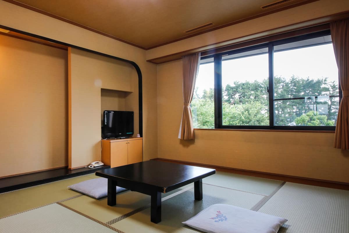 A traditional Japanese room with tatami flooring, a low table, a television on a cabinet, and a window with a view of trees.