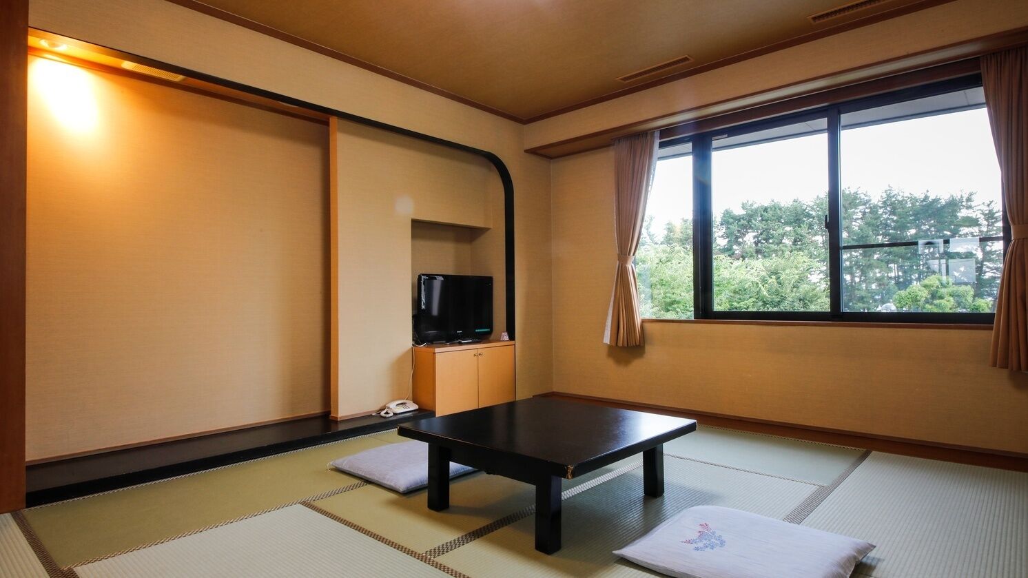 Una habitación con suelo de tatami, una mesa baja, un televisor sobre un mueble y una ventana con vistas a árboles.