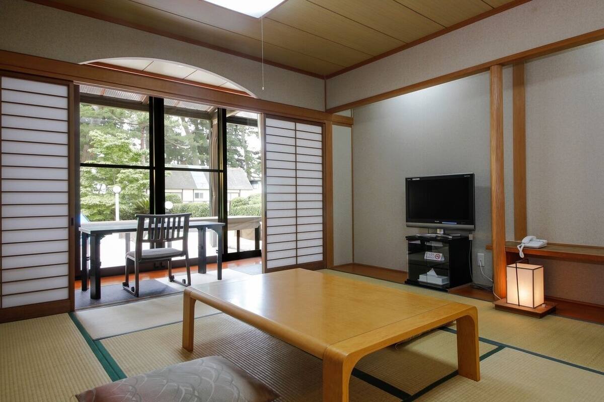 A traditional Japanese room with tatami flooring, a low wooden table, a television, and a sliding door leading to an outdoor area.