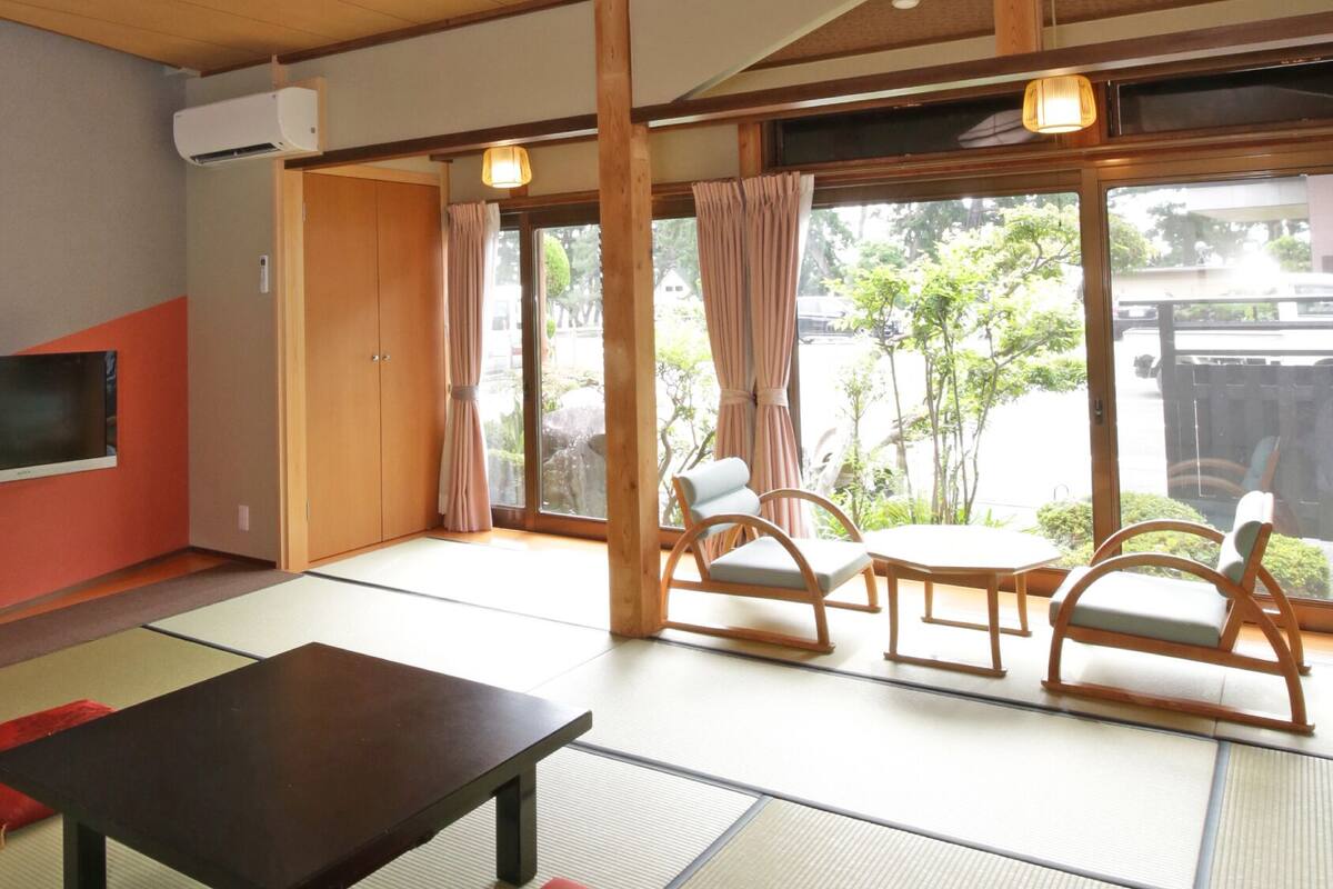 A room with tatami flooring, a television, a table, and a view of greenery outside.