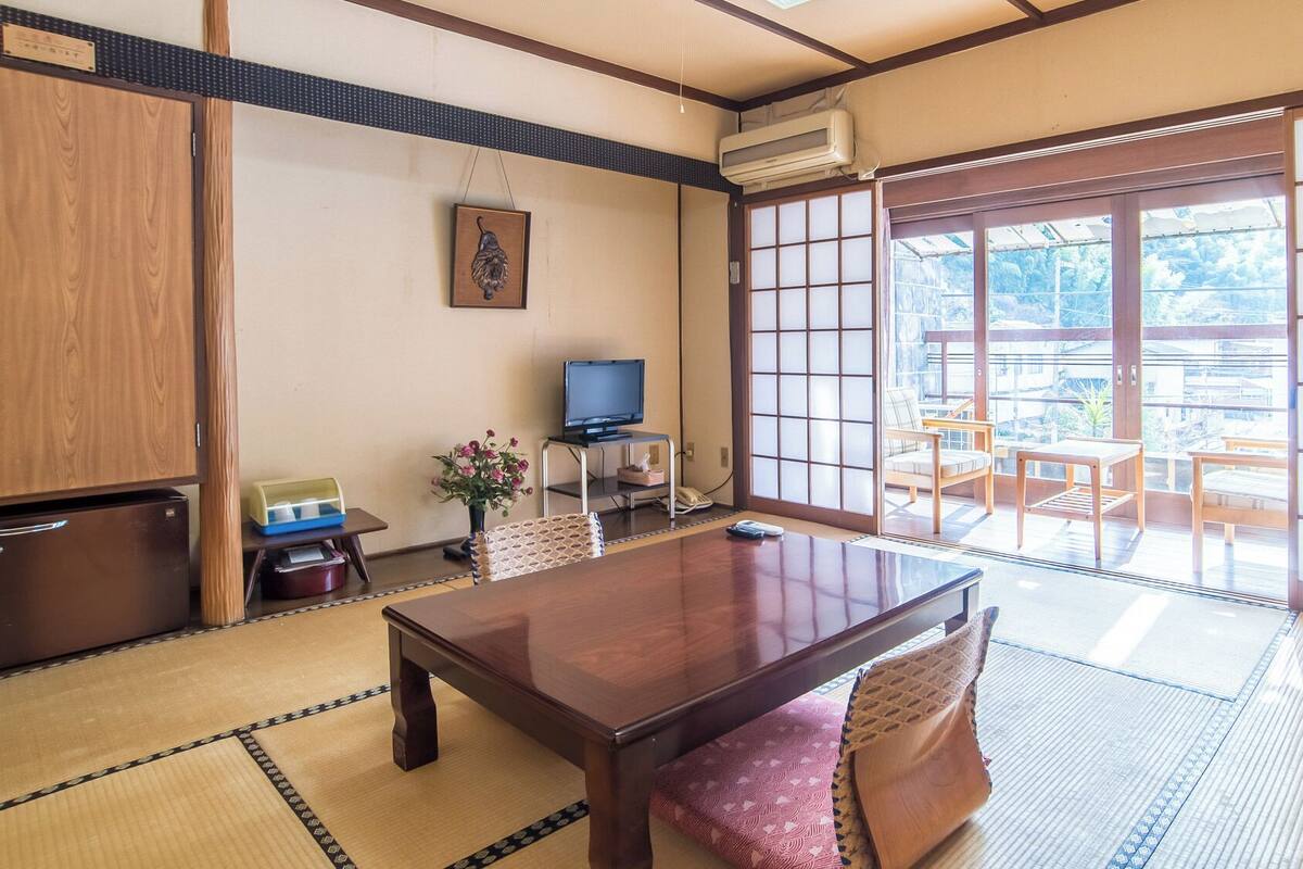 A traditional Japanese room with tatami flooring, a wooden table, a television, and a sliding door leading to an outdoor area.
