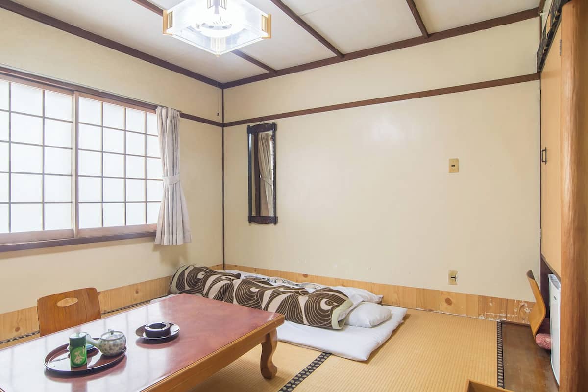 A traditional Japanese room with tatami flooring, a low wooden table with tea set, a cushioned bench, and a large window.