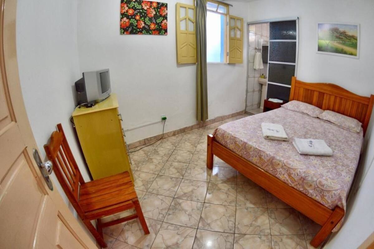 A bedroom with a bed, a wooden chair, a small desk with a television, and a bathroom with a mirror and sink.