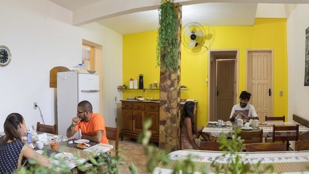 A dining area with a yellow wall, a fan, and a painting. - Pousada e Hostel Chapada Suítes (Lençóis)