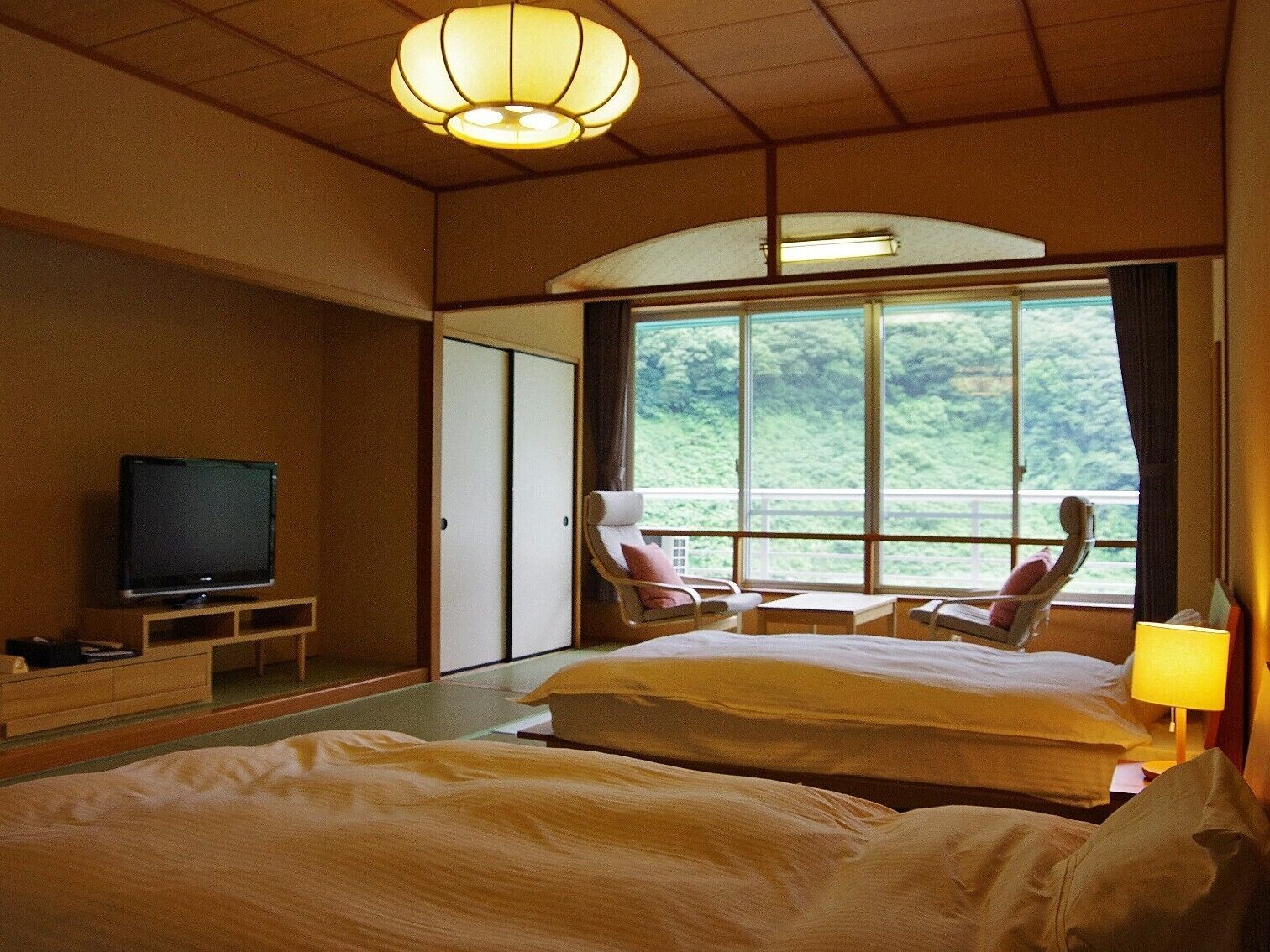 Een hotelkamer met een bed, een televisie, een stoel en een groot raam met uitzicht op groen.
