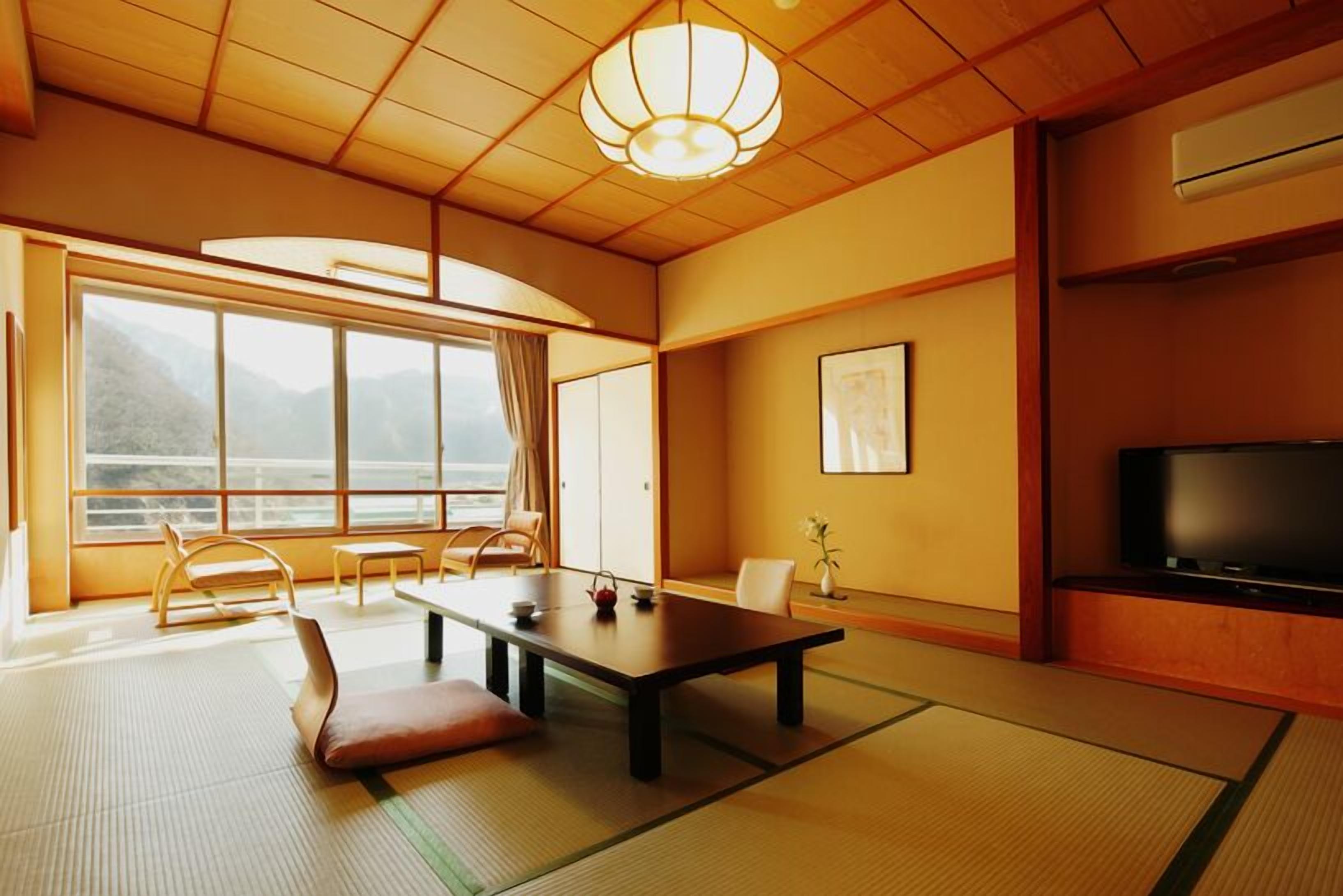 Een traditionele Japanse kamer met tatami vloerbedekking, een lage tafel, stoelen, een televisie en een groot raam met uitzicht op bergen.