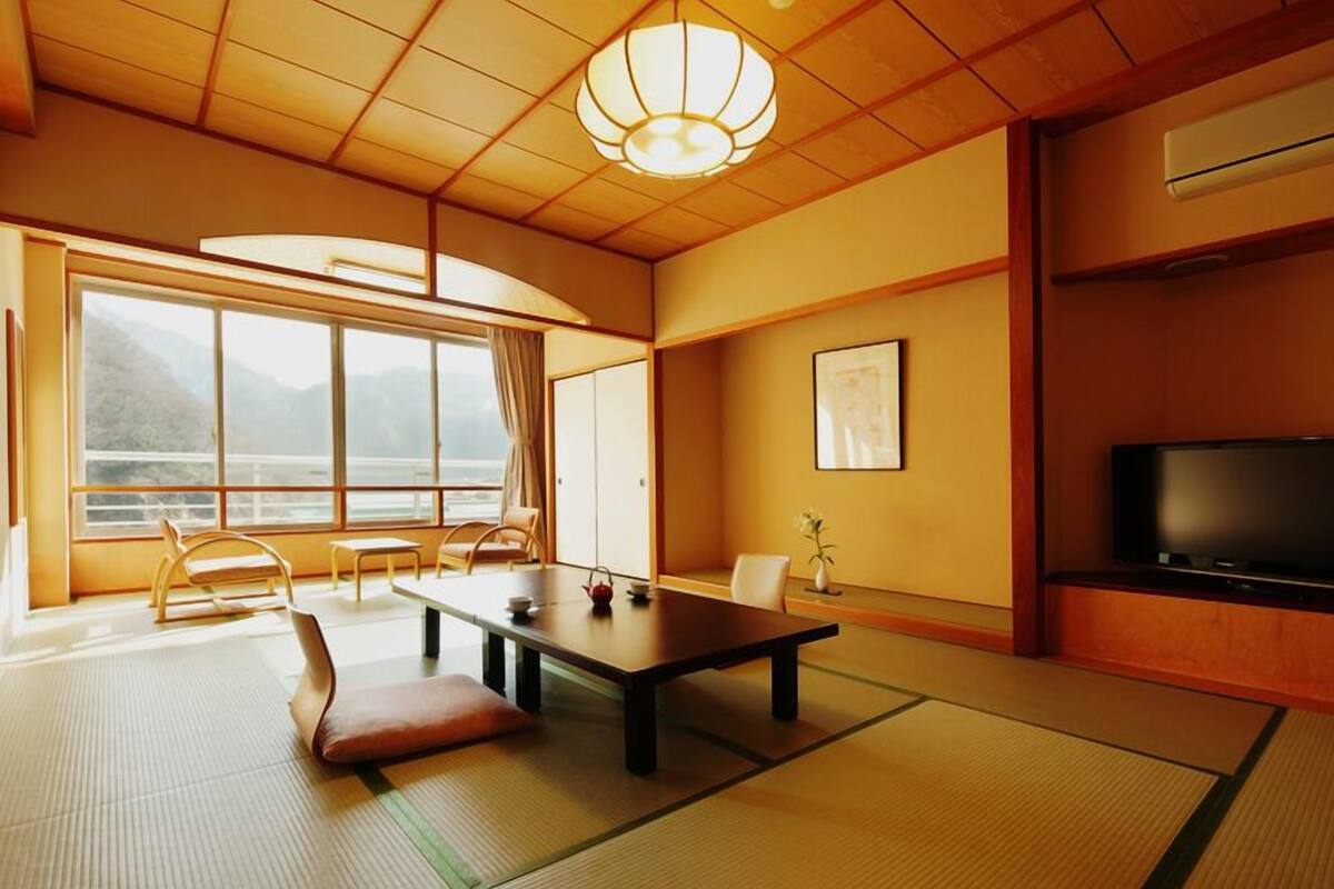 Una habitación japonesa tradicional con suelo de tatami, una mesa baja, sillas, un televisor y un ventanal con vistas a montañas.