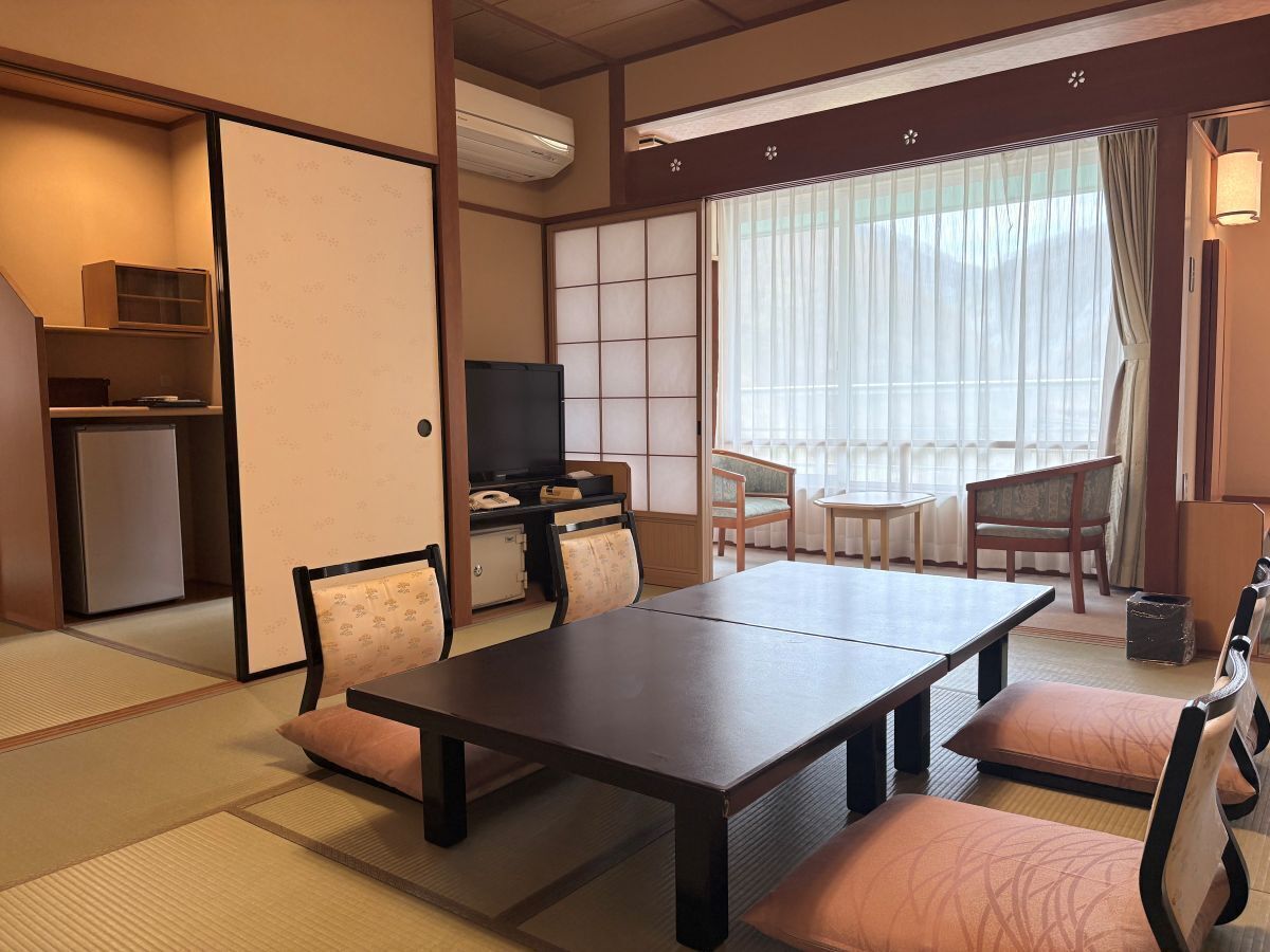 Ein traditioneller japanischer Raum mit Tatami-Böden, einem niedrigen Tisch, gepolsterten Sitzgelegenheiten, einem Fernseher und Schiebetüren.