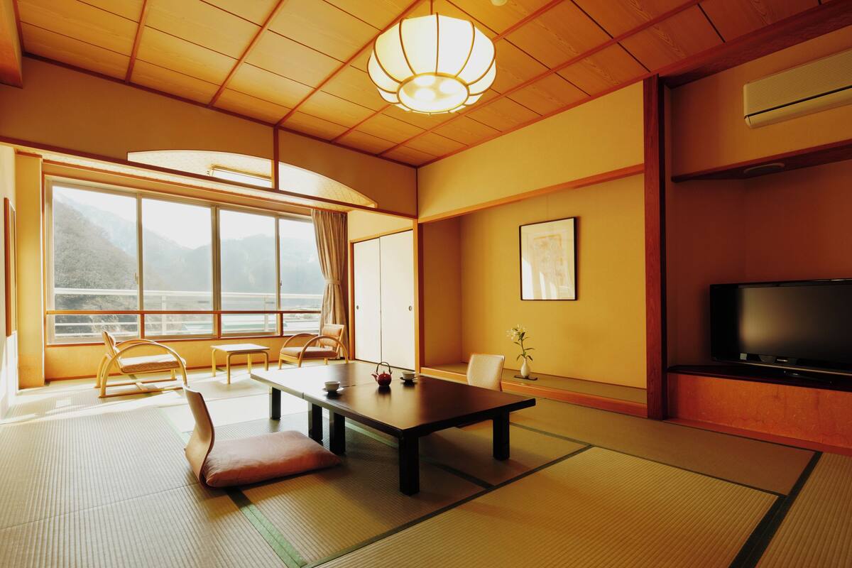 Una habitación japonesa tradicional con suelo de tatami, una mesa baja, sillas, televisión y vistas a montañas a través de amplios ventanales.