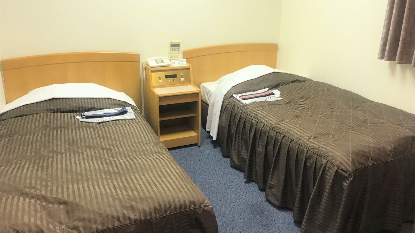 A hotel room with two single beds, a bedside table with a phone, and a small shelf.
