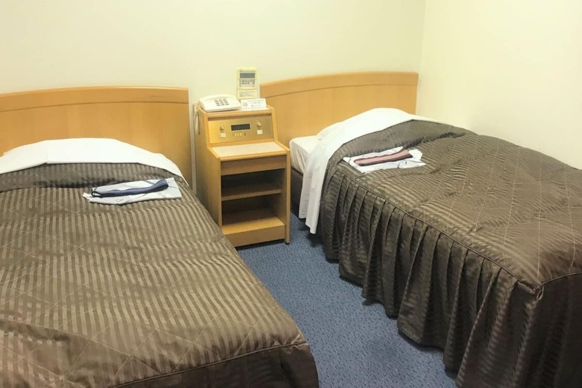 A hotel room with two single beds, a bedside table with a phone, and a small shelf.