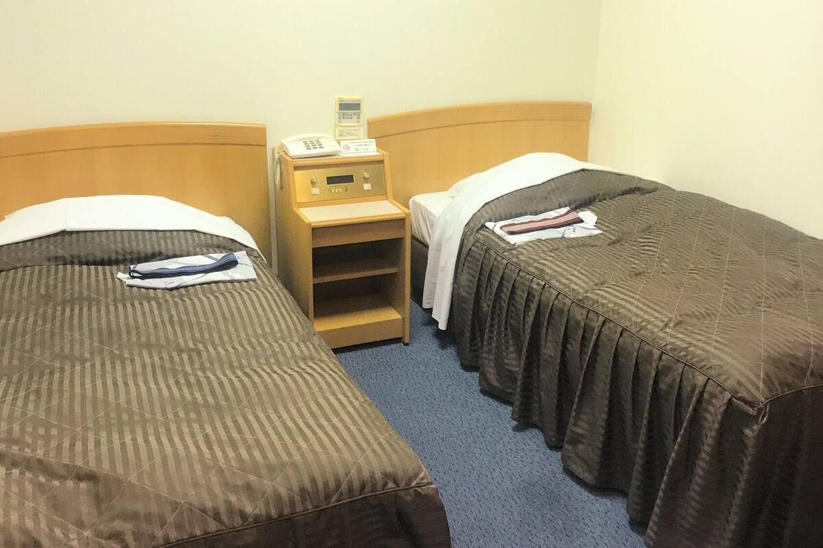A hotel room with two single beds, a bedside table with a phone, and a small shelf.