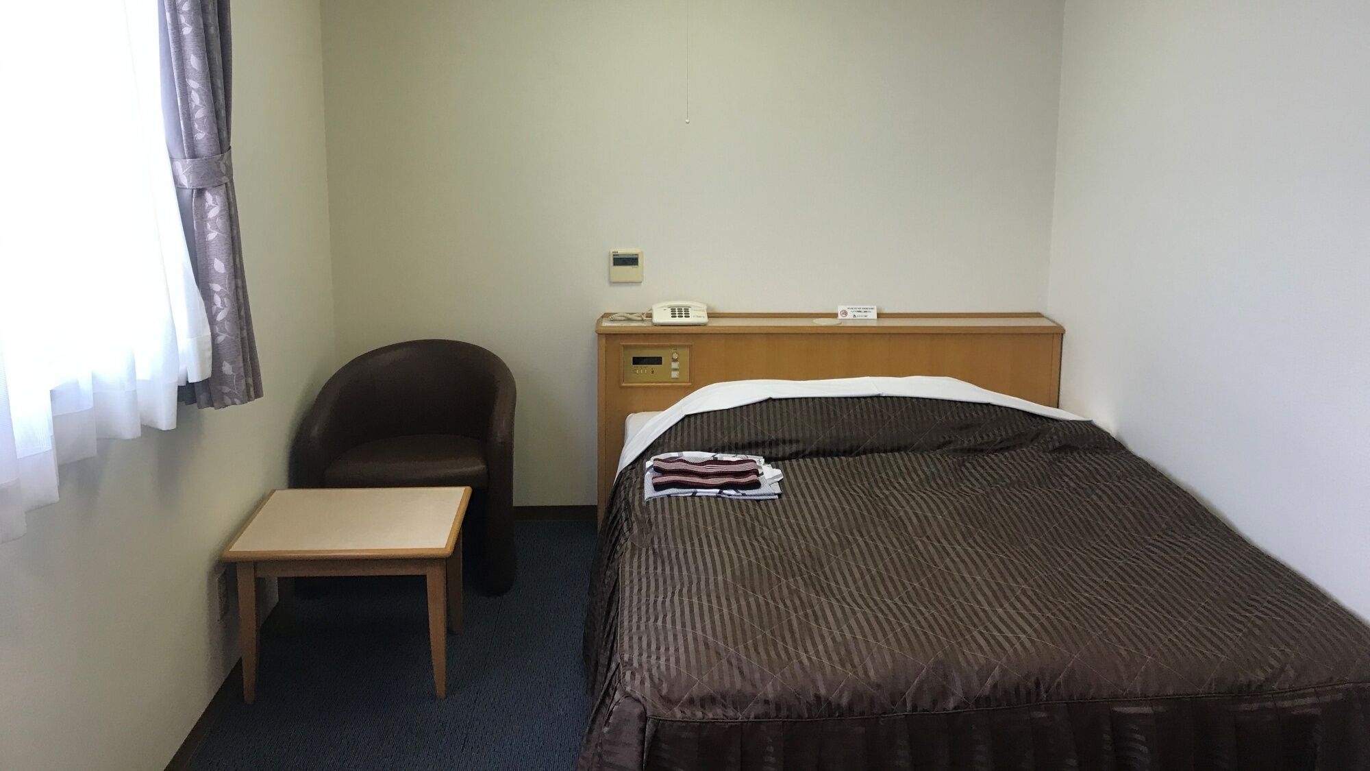 A small hotel room with a single bed, a wooden nightstand, a chair, and a small table.