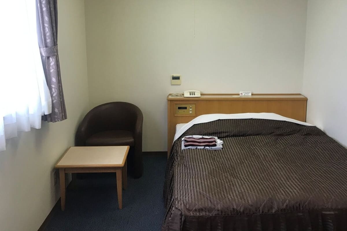 A hotel room with a single bed, a small wooden table, a chair, a bedside telephone, and a wall-mounted control panel.