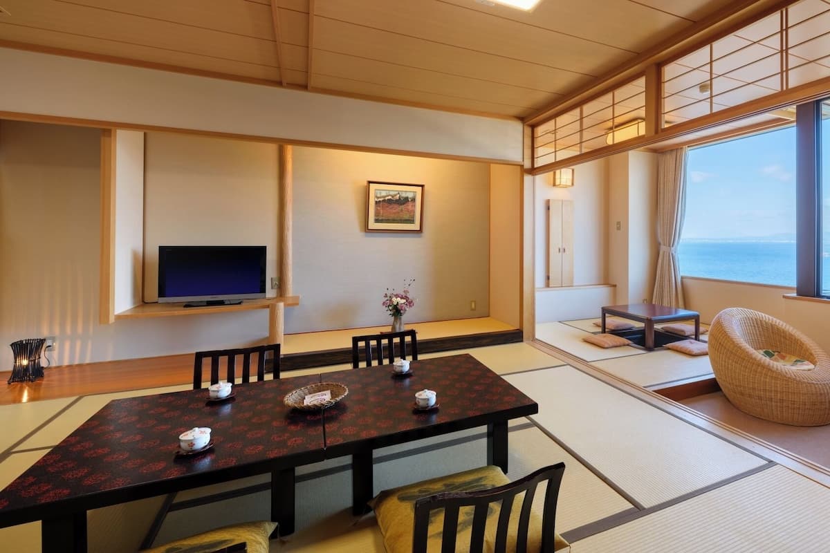 Ein traditionell japanisch gestalteter Raum mit Tatami-Böden, einem niedrigen Tisch mit Kissen, einem Fernseher und Blick auf das Meer.