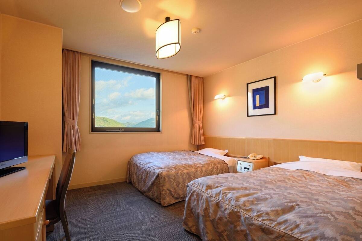 Ein Hotelzimmer mit zwei Betten, einem Schreibtisch mit Fernseher, einem Fenster mit Blick zum Himmel und einer Wandleuchte.
