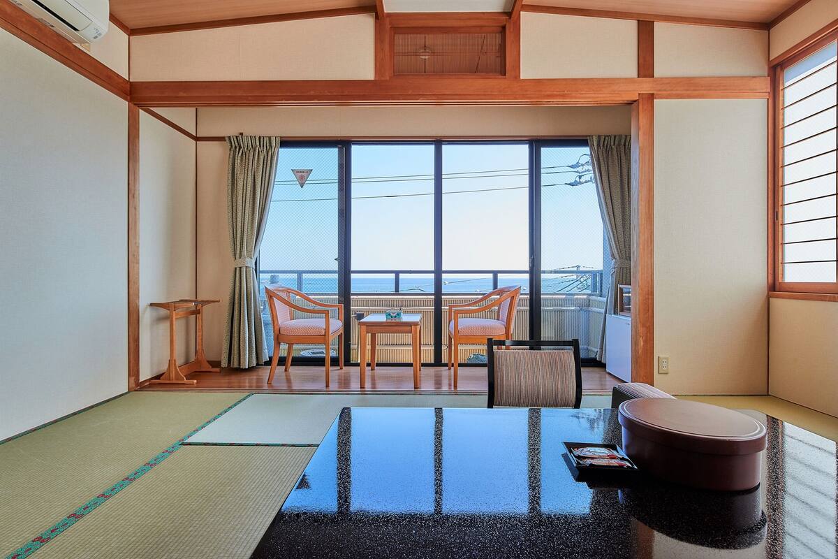 A traditional Japanese room with tatami flooring, a low table, two chairs, and a view of the sea through large windows.