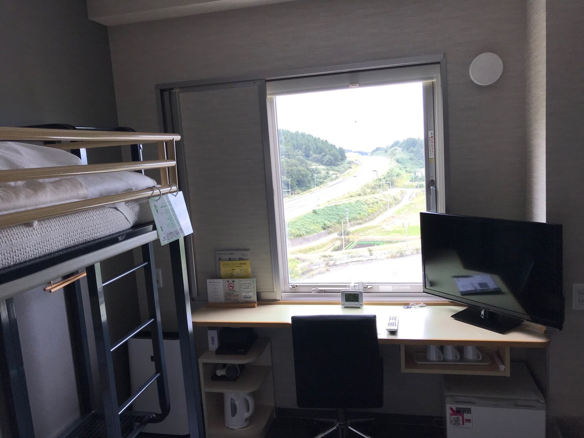 A bunk bed, a desk with a computer, a TV, and a window with a view of a road and hills.