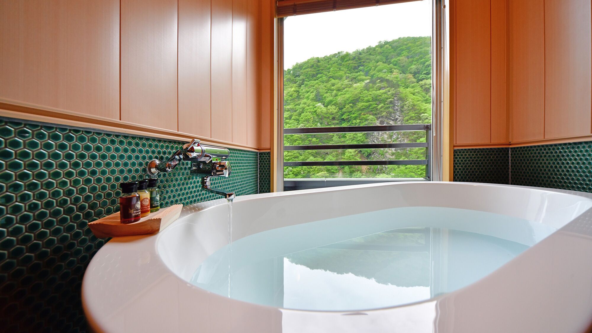 Une salle de bain dotée d’une grande baignoire, d’un pare-feu en carreaux verts, de meubles en bois et offrant une vue sur une forêt par la fenêtre.