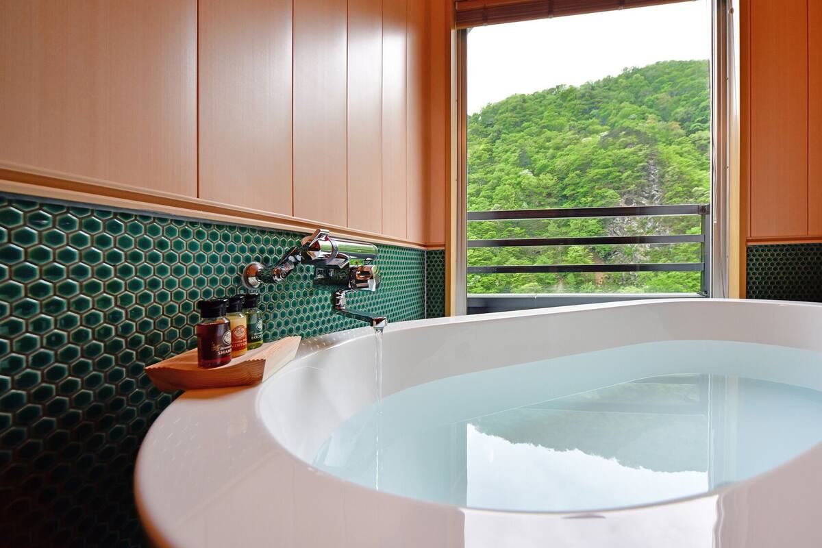 Une salle de bain dotée d’une grande baignoire, d’un pare-feu en carreaux verts, de meubles en bois et offrant une vue sur une forêt par la fenêtre.