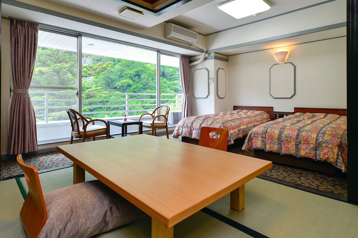 Une chambre d’hôtel avec une table en bois, deux lits avec une literie à motifs floraux, une vue sur la verdure grâce à de grandes fenêtres et un ventilateur de plafond.