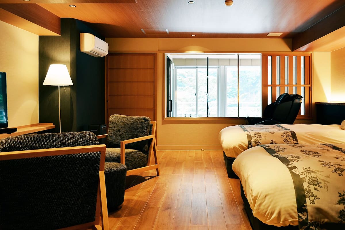Une chambre d’hôtel avec deux lits, un bureau avec une chaise, une lampe et une fenêtre avec des stores.